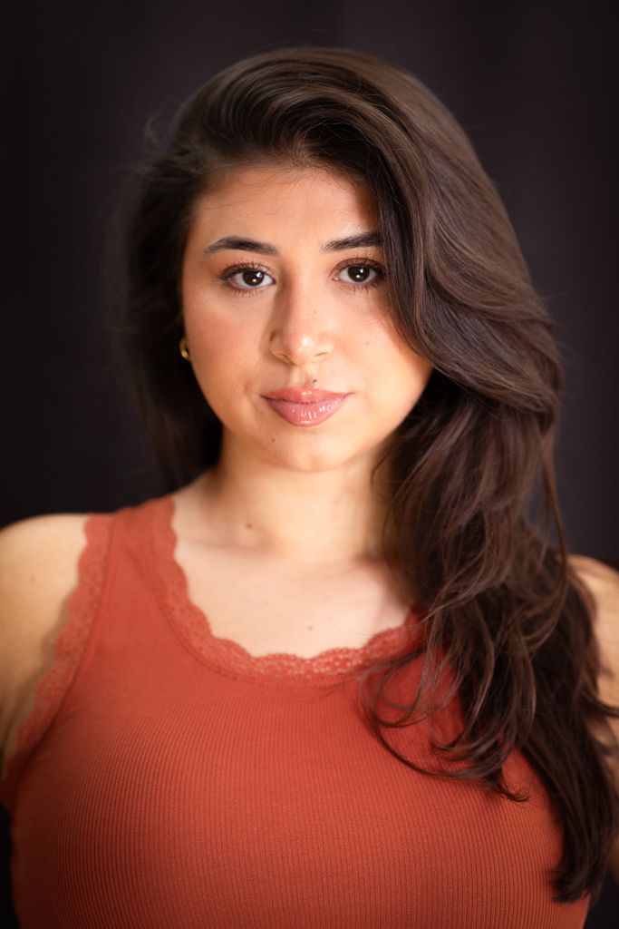 Close-up portrait of a woman with long brown hair in a rust tank top against a dark background.